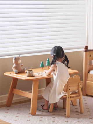 Child sitting at a wooden table with toys in a room with a window and bed.