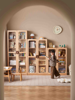 Child standing in front of a wooden bookshelf filled with toys and books in a room with a clock on the wall.