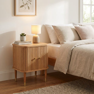 Wooden nightstand with lamp and books next to a bed in a bedroom.